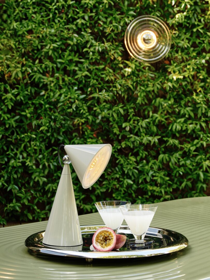 A small table lamp and two cocktail glasses sit on a silver tray with a halved passion fruit. A green leafy background is visible behind