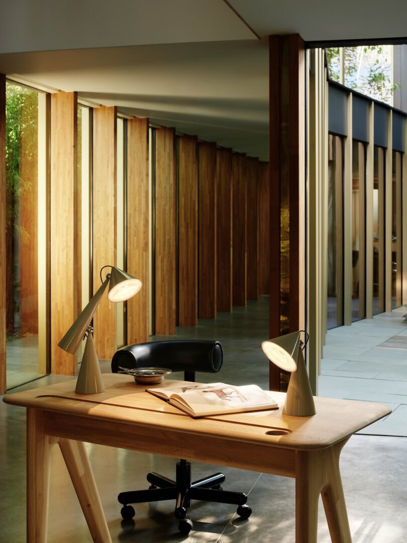 Modern office interior with a wooden desk, open book, two angled lamps, a black chair, and large windows showing wooden paneling and greenery outside