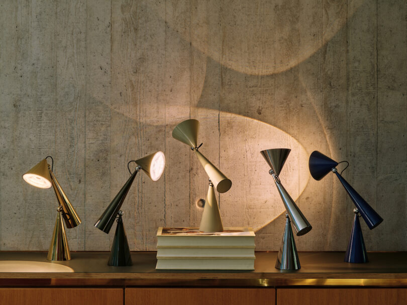 A row of five cone-shaped desk lamps in various colors, positioned creatively on a wooden surface with books, casting light on a textured wall