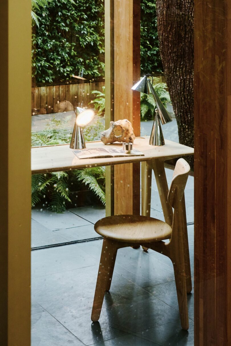 A wooden desk with two lamps and a skull on it is positioned next to a large window overlooking a green garden. A matching wooden chair is placed in front of the desk