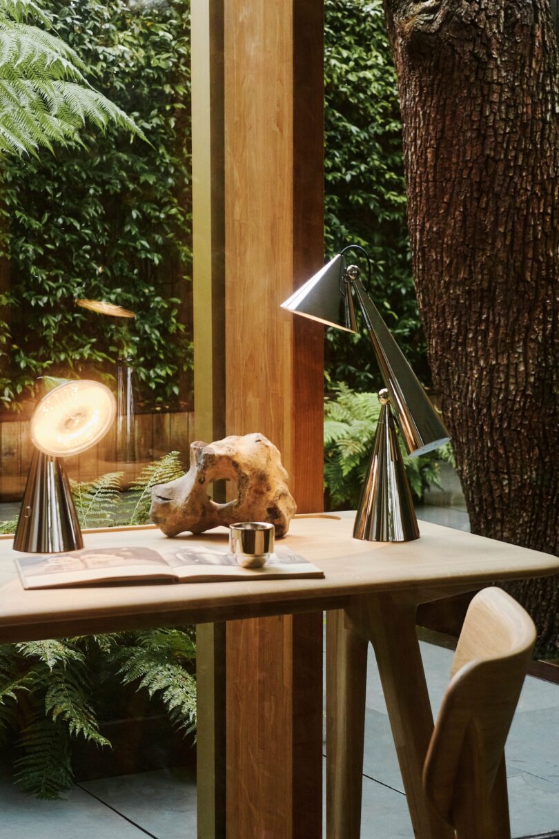 A wooden desk with a conical lamp, a sculptural rock, a small candle holder, and an open book. It is near glass windows overlooking lush greenery and a tree trunk