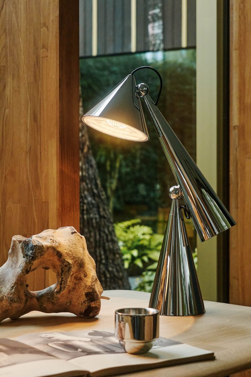 Modern metallic desk lamp illuminated on a table with a rock, a small cup, and an open book. Background shows a window with greenery outside