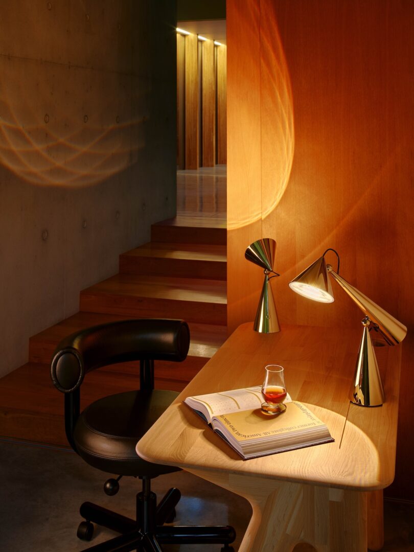 A warm, softly lit room with a wooden desk, modern lamp, black chair, open book, and a glass of amber liquid. Steps and a concrete wall are visible in the background