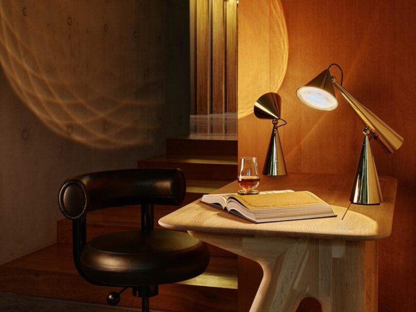 A modern desk with an open book, a glass of whiskey, and two angular lamps. A black chair sits nearby, and warm lighting creates a cozy atmosphere