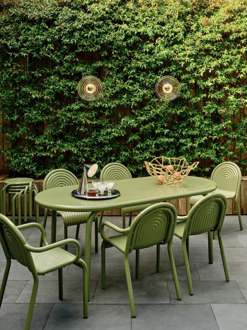 Green patio set with oval table and eight chairs on a tiled patio. A tray with a pitcher and glasses sits on the table. Lush vertical garden and warm lighting in the background