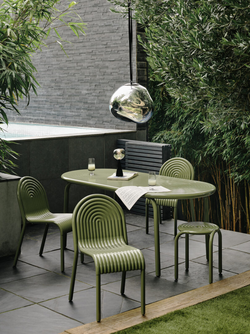 Outdoor dining set with green ribbed chairs and table on a stone patio, surrounded by lush bamboo. A metallic pendant light hangs above, and two glasses with water are on the table