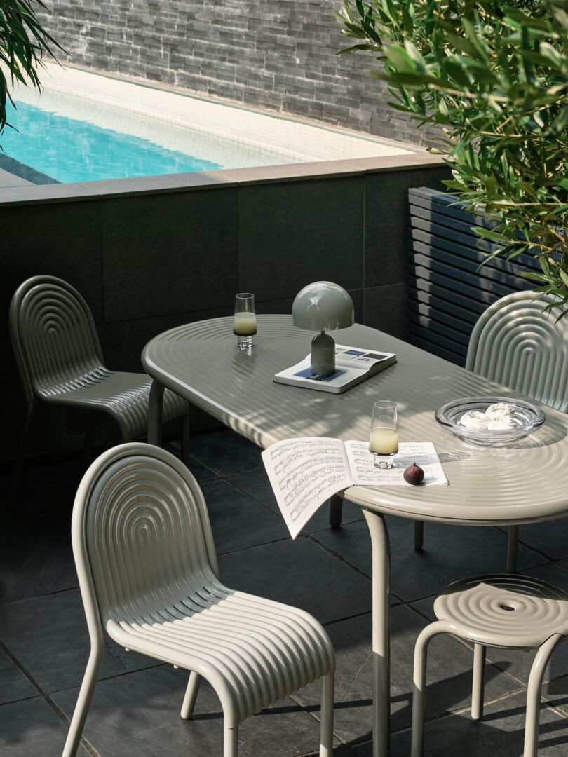 Elegant outdoor patio with a curved table, four chairs, and a lamp. Two drinks, an open magazine, and a bowl with snacks are on the table. Pool in the background with greenery around