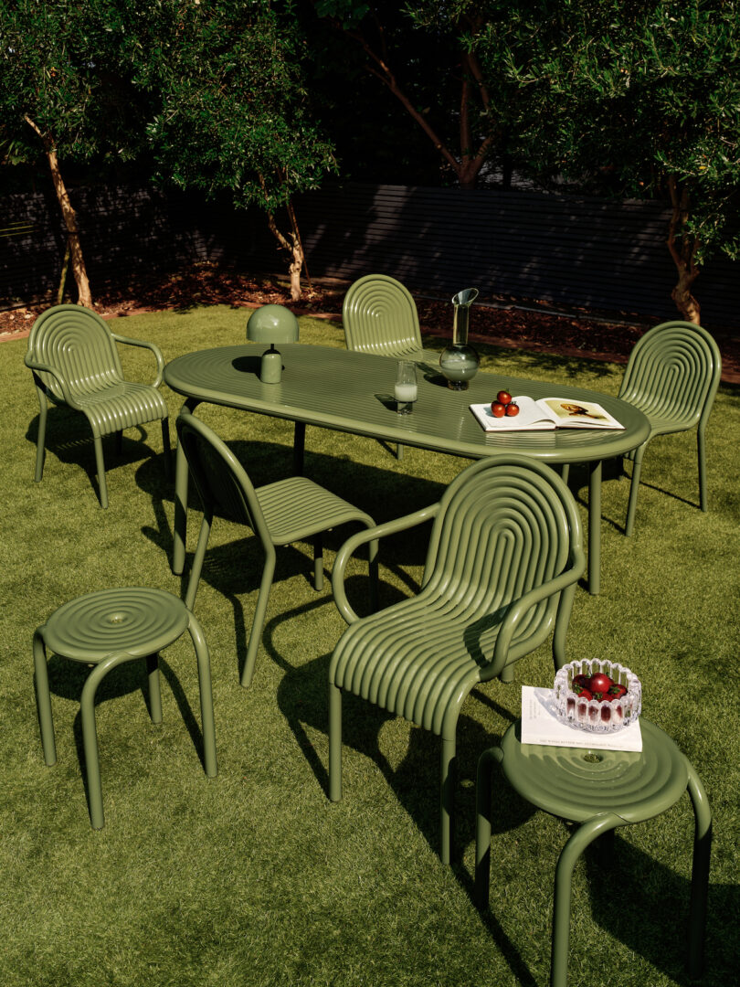 Outdoor dining setup with green chairs and a matching table on grass. The table holds glassware, a carafe, and a dish with fruit. Chairs display a ribbed design. Trees and a black fence in background