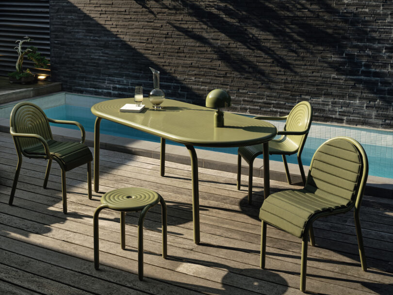 Outdoor patio with green metal furniture, including a table, four chairs, and a stool beside a pool. A decanter, two glasses, and a small notebook are on the table