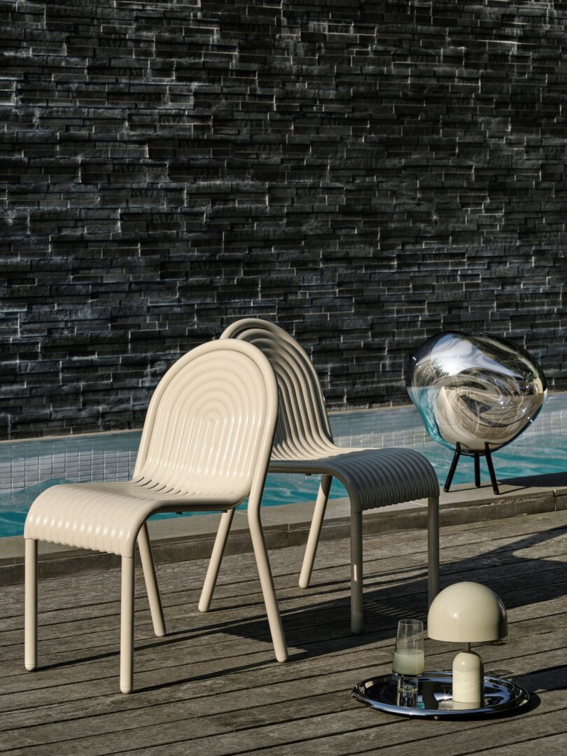 Two beige chairs on a wooden deck beside a pool, with a spherical decorative object and a tray with a candle and glass. Stone wall backdrop