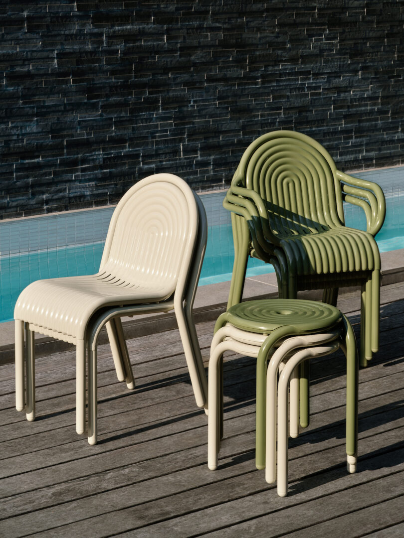 Four modern, ribbed chairs and two stacked stools in beige and green are arranged on a wooden deck near a swimming pool with a dark stone wall backdrop