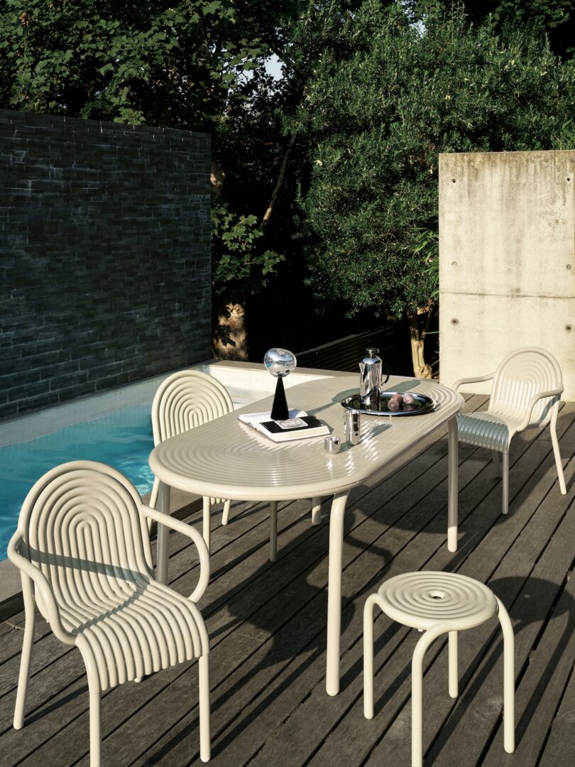 Outdoor patio with a cream-colored table set, four chairs, and a stool on a wooden deck beside a pool. Trees and a stone wall in the background. Items on the table include a book and a globe