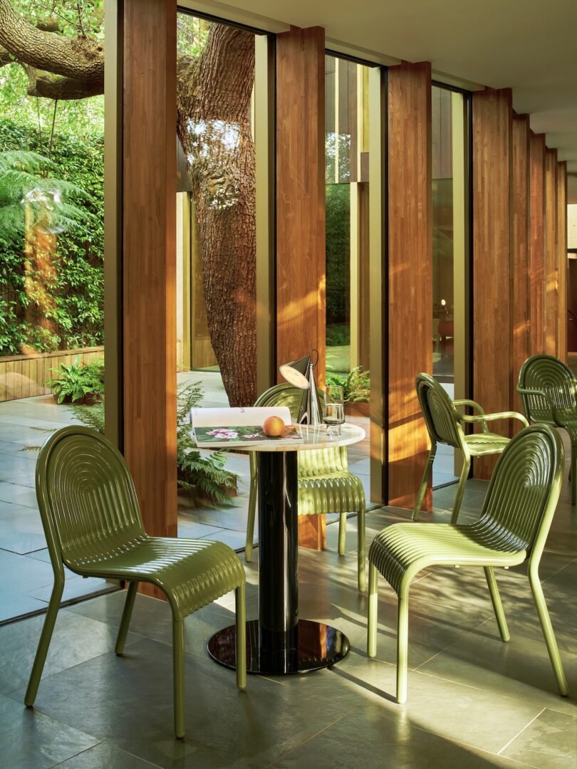 A modern dining area with green chairs and a round table, next to large windows revealing a tree and garden outside