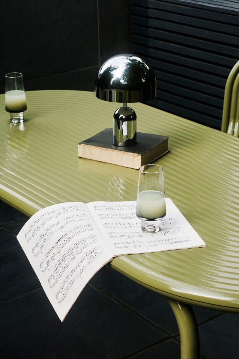 Sheet music and a glass of green liquid on a table with a lamp and two books. Another similar glass is on the table