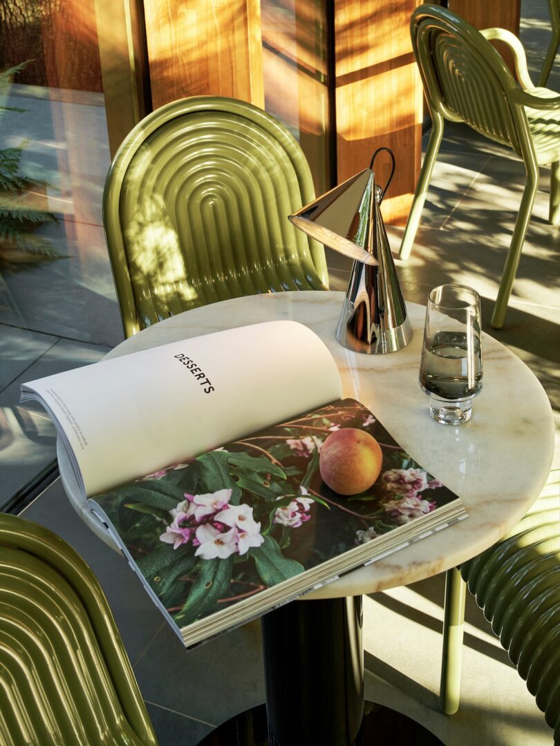 A small round table with an open book showing floral images, a water glass, and a metallic cone-shaped lamp. Green chairs and sunlight filtering through windows in the background