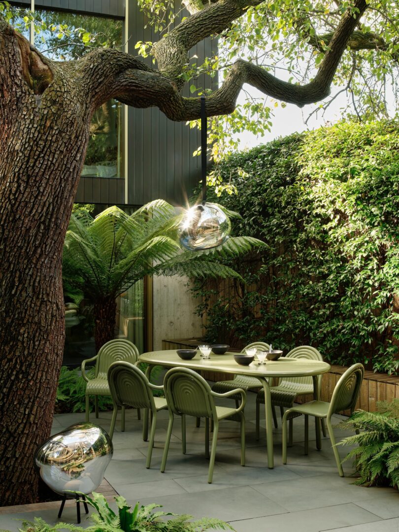 Outdoor dining area with green chairs and table under a large tree. Hanging glass lamps and lush greenery surround the space, creating a tranquil garden setting