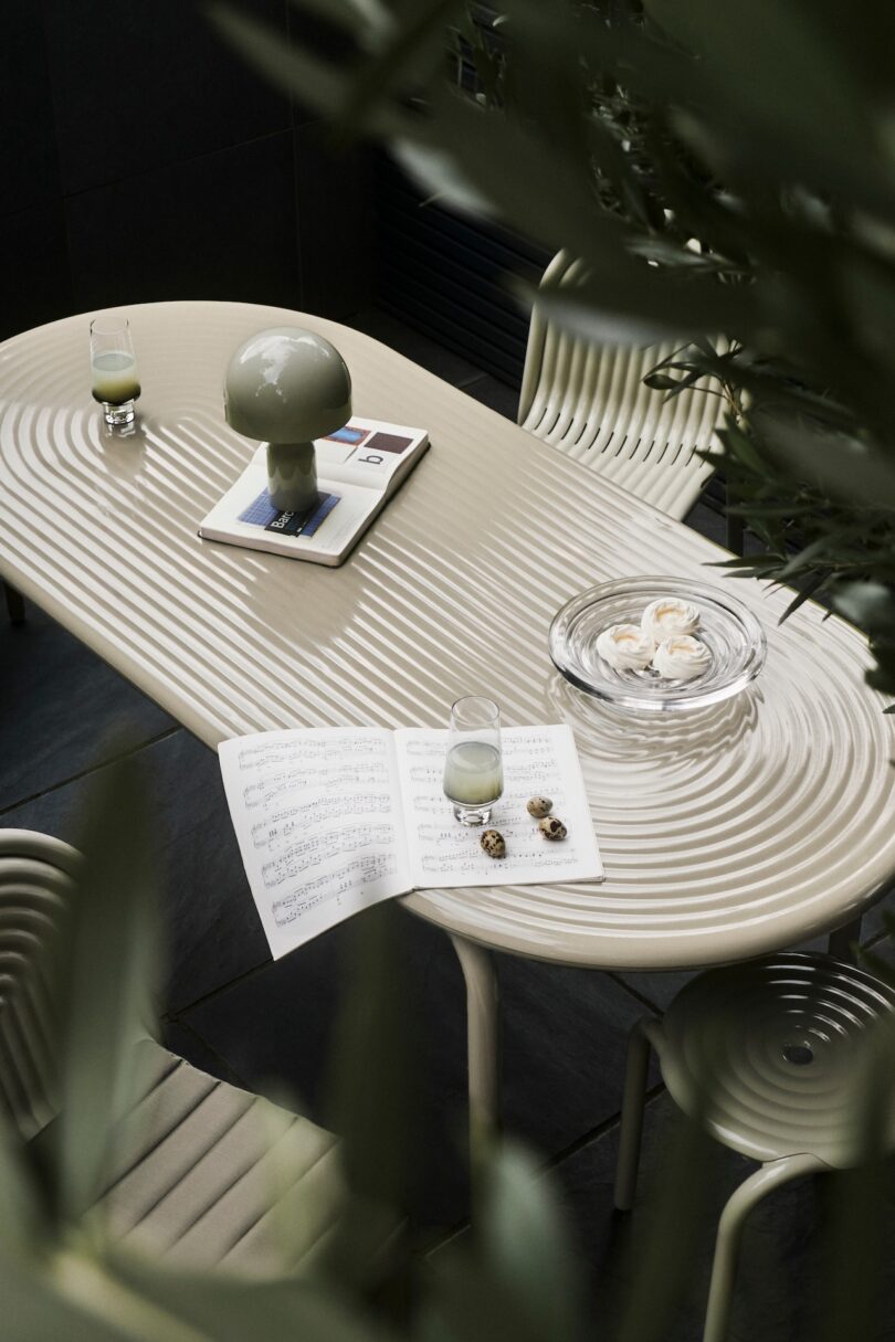 Oval table with a ripple design, two glasses, a lamp on a book, a glass dish with seashells, sheet music, and green chairs. Surrounded by foliage