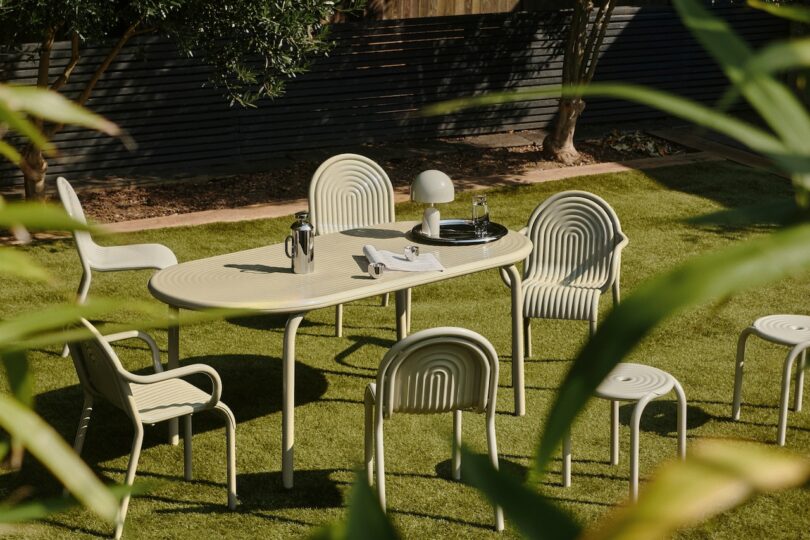 Outdoor dining area with a cream-colored table and chairs on grass. There's a lamp and cups on the table. Surrounded by plants