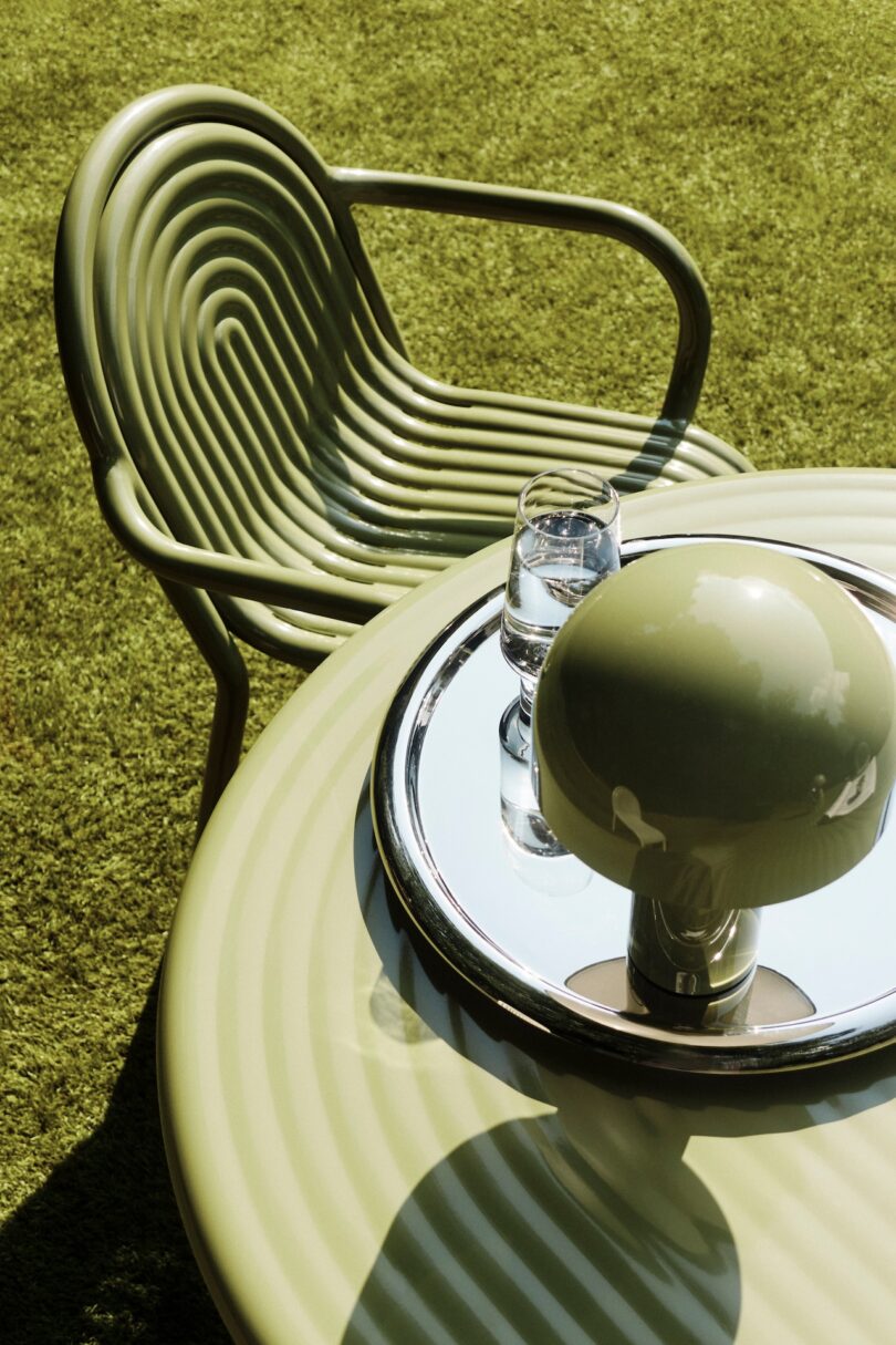 Green metal chair next to a matching round table with a wavy design. On the table, there's a glass of water and a circular lamp on a mirrored tray, set against a grassy background