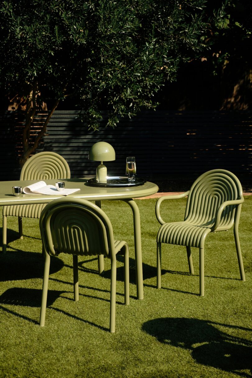 Outdoor setting with a round table, three green chairs, a lamp, and a glass of water on grass, surrounded by greenery and a dark fence