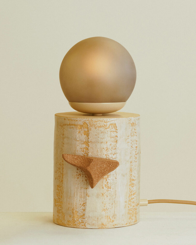 A spherical light fixture rests atop a wooden cylindrical base with a triangular cork accent, set against a neutral background