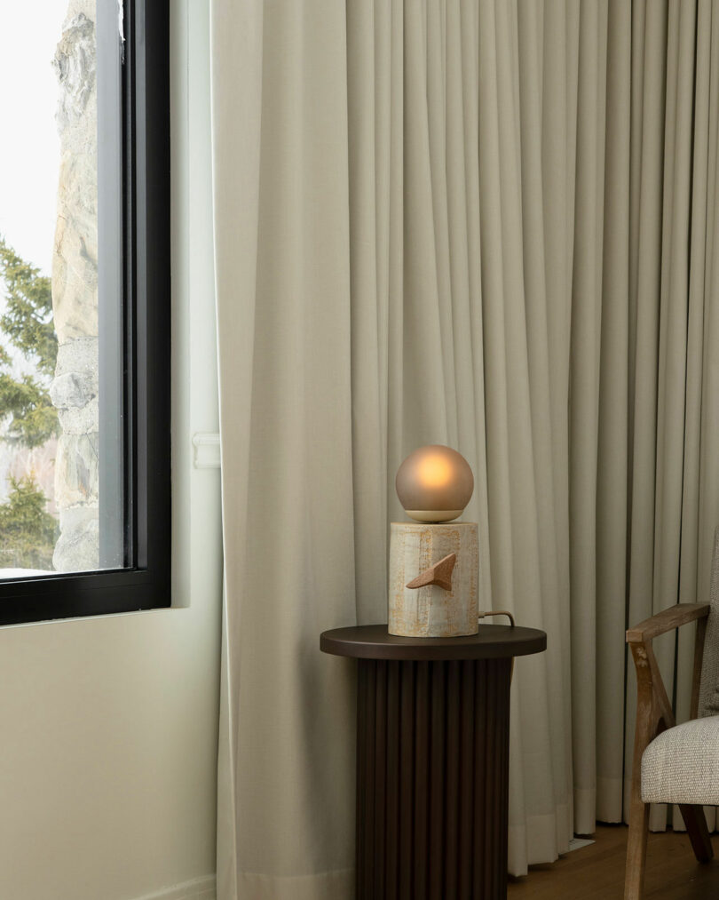 A small lamp with a round bulb on a dark side table next to a wooden chair, set in a room with beige curtains and a window