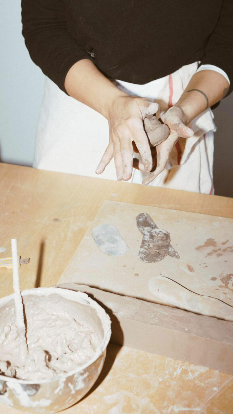 A person in a black shirt and white apron molds clay with their hands at a table, next to a bowl of wet clay and a clay-covered wooden board