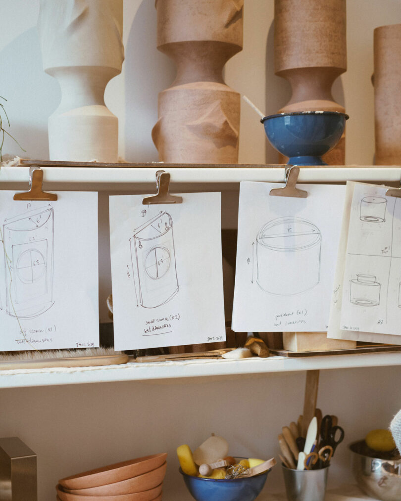 Sketches of pottery designs clipped on a shelf, with vases and bowls in the background