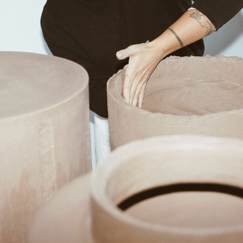 Person shaping a large, round clay sculpture with one hand, wearing a black top with visible tattoos on the arm. Another large clay piece is nearby