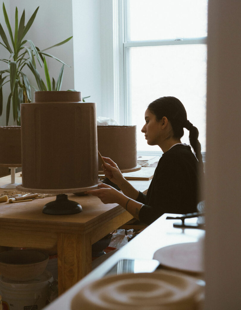 Person focused on sculpting a large cake in a kitchen, with tools and plants nearby