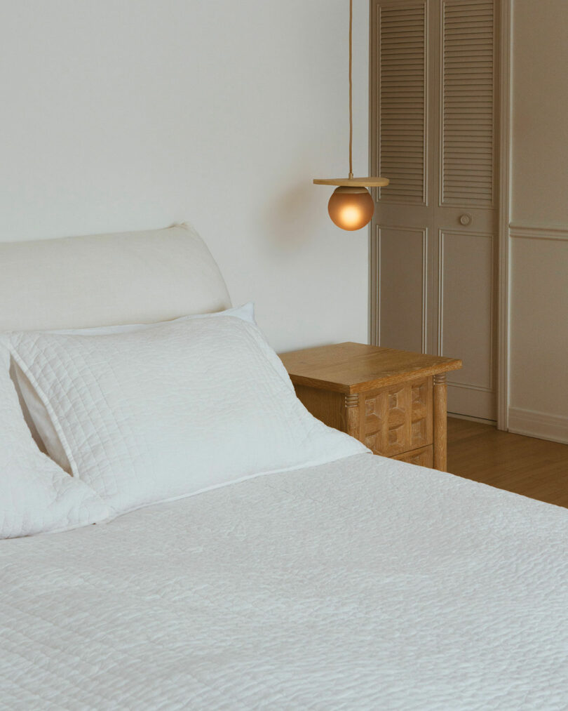 Minimalist bedroom with a white bed, wooden nightstand, and a small hanging lamp. Closed closet doors in the background