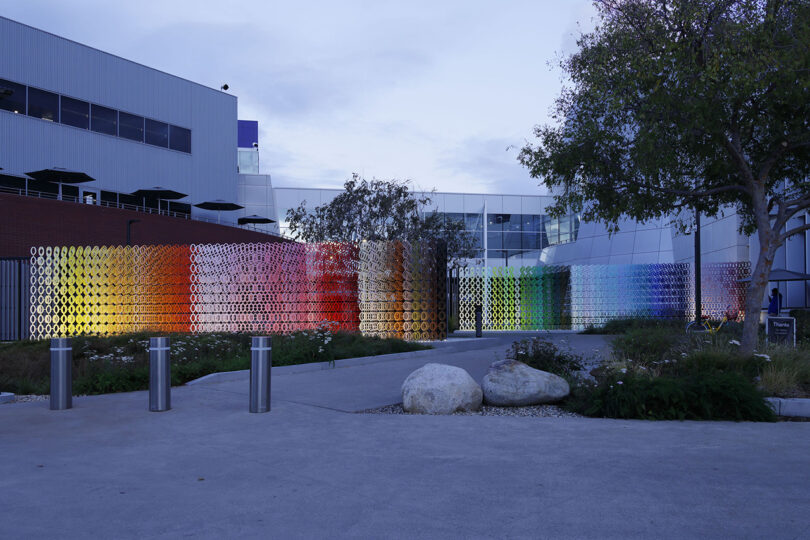 A colorful, circular-patterned wall installation outside a modern building, with trees and greenery nearby.