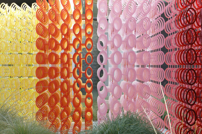 Colorful art installation featuring vertically arranged rings in yellow, orange, and pink, creating a gradient effect. Green plants are visible at the bottom of the image.