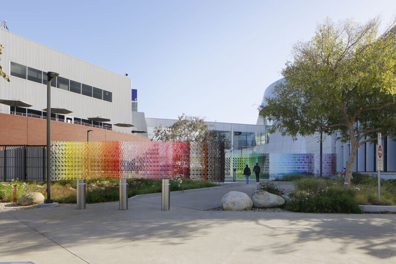 Colorful circular art installation in a courtyard between modern buildings, with a tree and two people walking nearby.