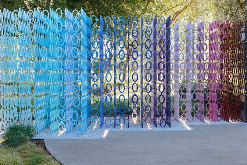 Vertical panels of intertwined circular patterns in gradient colors from blue to purple create a stunning display, reminiscent of the vibrant works by 100 Colors Emmanuelle Moureaux, set gracefully in an outdoor park with trees and a concrete path.