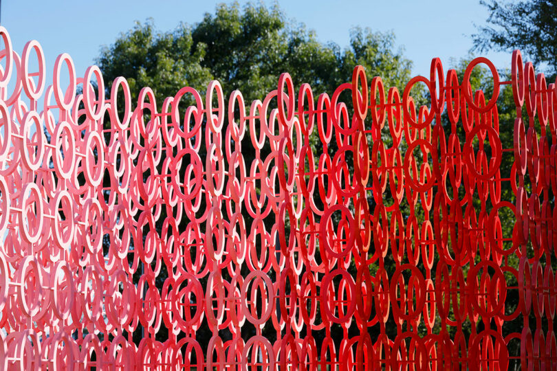 A red sculpture made of interlocking circular rings, reminiscent of Emmanuelle Moureaux's "100 Colors," is set against a backdrop of green trees and a clear blue sky.