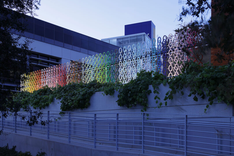 Colorful circular art installation on a building wall with greenery in front and a railing below.