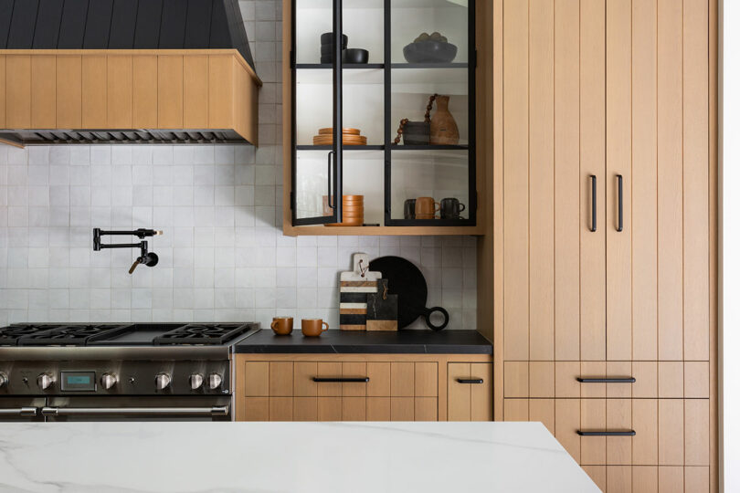 Cocina moderna con gabinetes de madera clara, una estufa de acero inoxidable, grifo negro y encimera blanca. Los gabinetes superiores tienen puertas de vidrio que muestran platos y artículos de decoración.