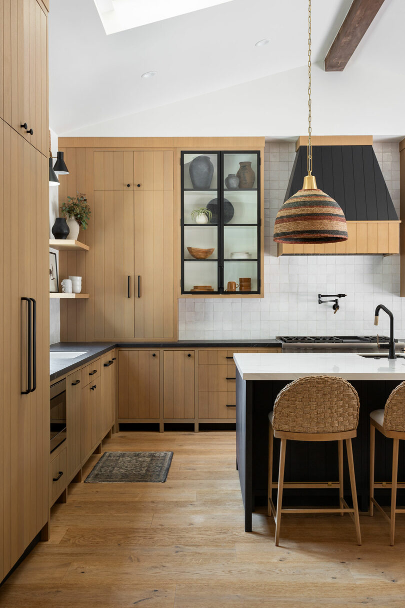 Cocina moderna con gabinetes de madera, isla con sillas tejidas, luz colgante y acentos oscuros. Los gabinetes muestran varios platos. La luz natural del tragaluz ilumina el espacio.