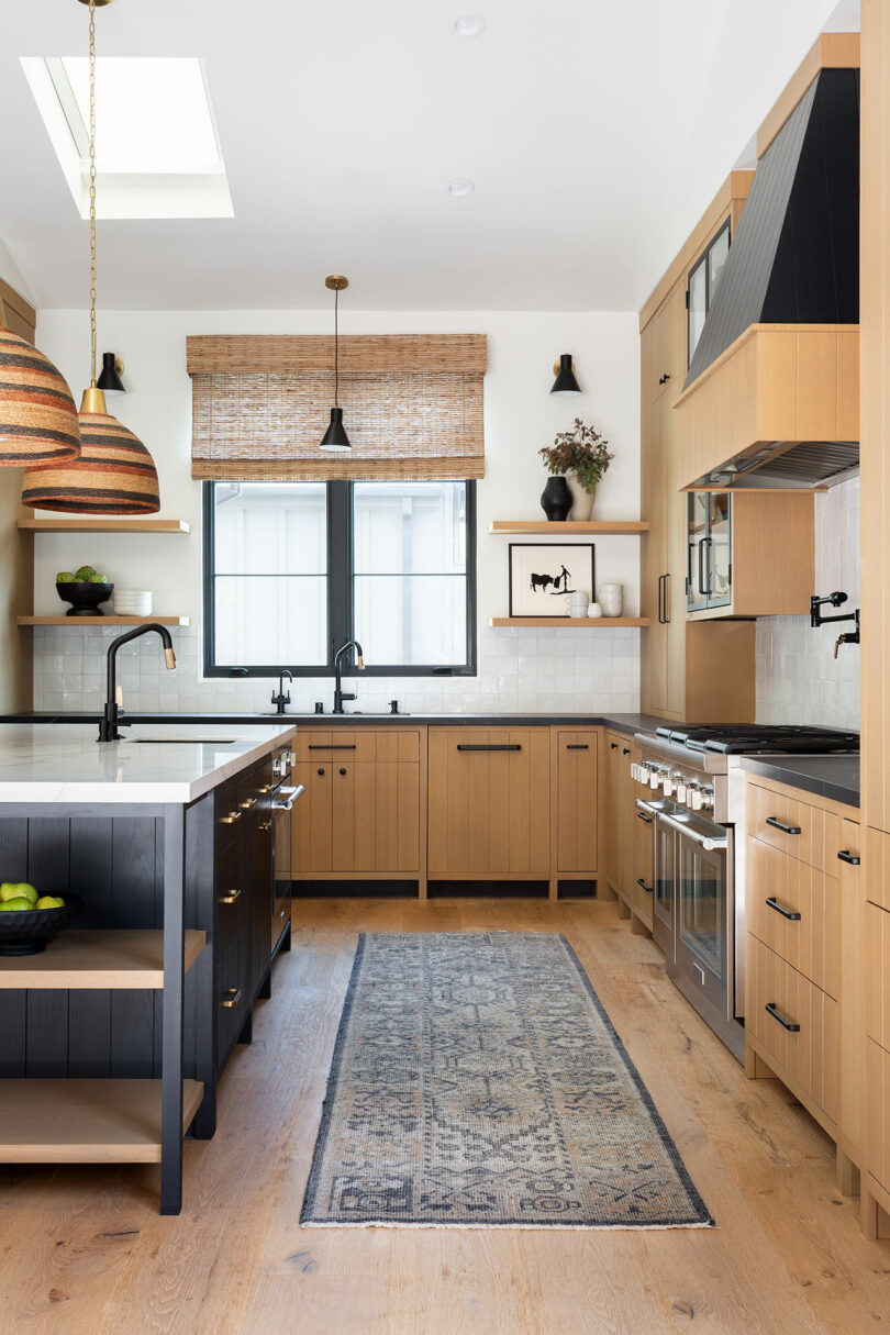 Una cocina moderna con gabinetes de madera, una isla central, una alfombra y dos luces colgantes. Una ventana con una ciega tejida y varios electrodomésticos de cocina es visible.