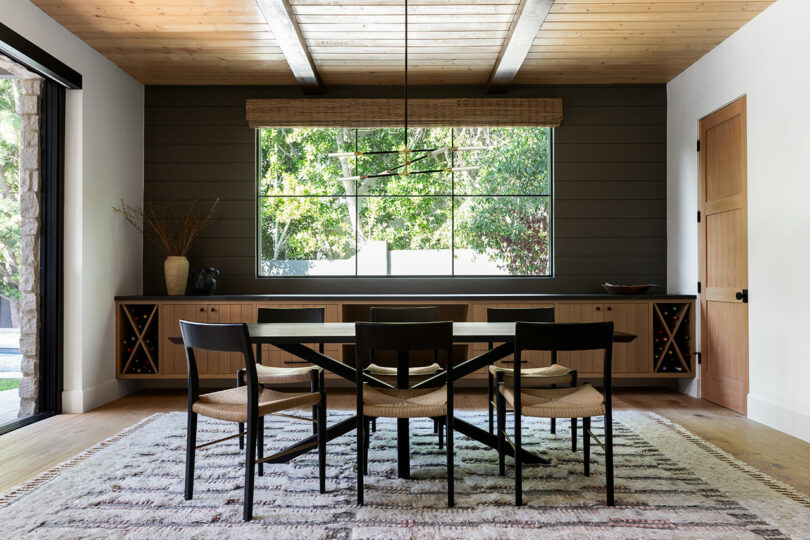 Comedor con una mesa de madera y seis sillas, una gran ventana con vista de vegetación, vigas de techo de madera, una alfombra tejida y dos estantes de vino en un aparador.