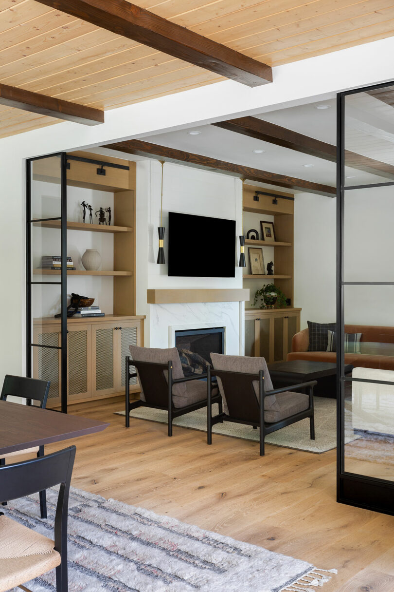 A modern living room with a wall-mounted TV above a fireplace, two chairs, a couch, and a dining table. Shelves with decor items and wooden beams accent the light wood flooring and ceiling.