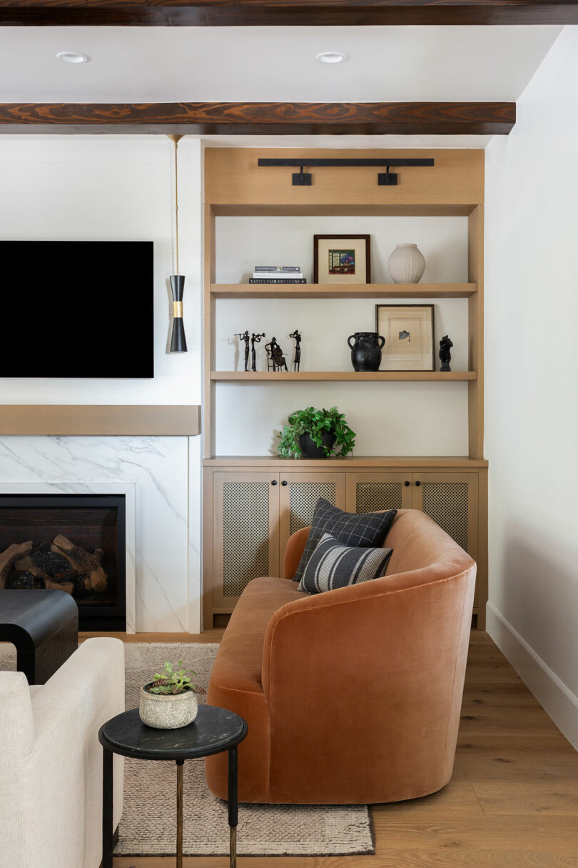 Sala de estar con un sofá curvo marrón, mesa lateral negra y estantes incorporados. Los estantes sostienen artículos decorativos y plantas. Una moderna chimenea y televisión montada en la pared están en la pared adyacente.