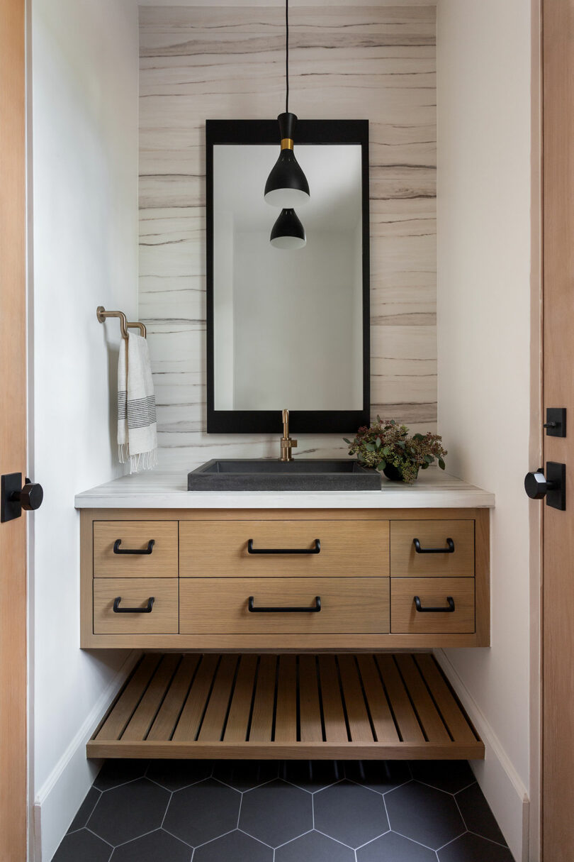 Baño moderno con tocador de madera, espejo de marco negro, fregadero negro y luz colgante minimalista. La pared blanca a rayas y las baldosas hexagonales del piso agregan contraste. Toalla en un soporte de latón.