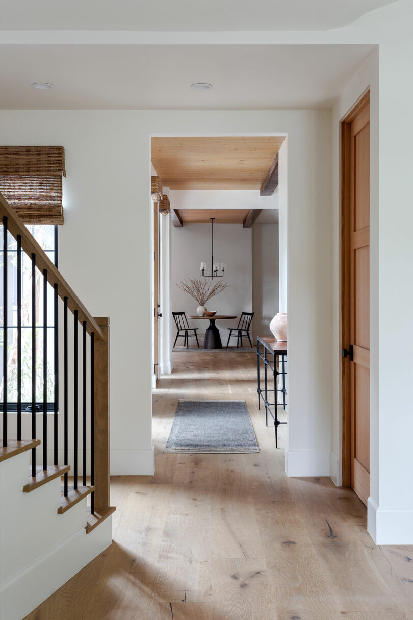 Pasillo interior con pisos de madera, una escalera a la izquierda y una mesa con un jarrón y ramas secas en el fondo. Dos sillas y una lámpara de araña son visibles en el comedor.