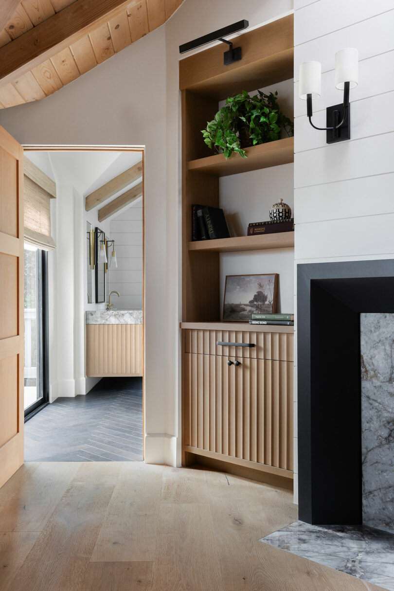 Interior view of a room with wooden flooring, a fireplace, and built-in shelving. There is a doorway leading to a bright room with a sink and mirror. A potted plant is on the shelf.