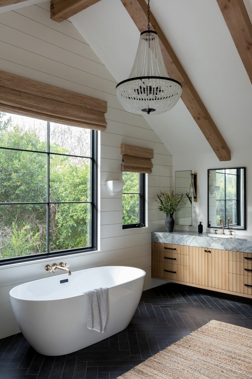 Un baño moderno con una bañera independiente, ventana grande, tocador de madera con encimera de mármol y una lámpara de araña. El piso de la espiga oscura y la luz natural completan la escena.