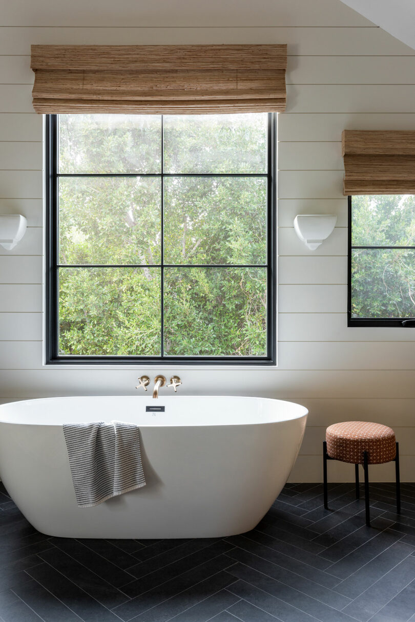 Un baño moderno con una bañera independiente, una toalla gris cubierta sobre el costado, una ventana con una vista frondosa, un pequeño taburete y dos luces de pared.