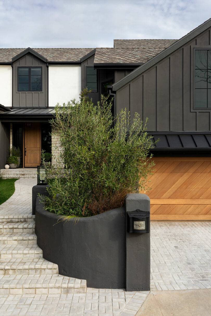 Modern two-story house with dark gray and white exterior, wooden garage door, and curved gray wall. Pathway leads to the entrance beside the garage. Small tree in the front garden.