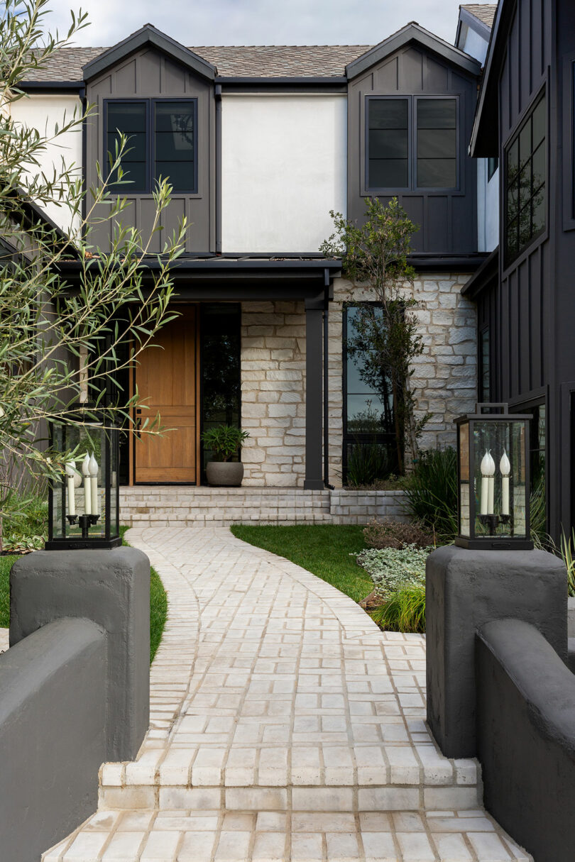 Exterior moderno de la casa con acentos oscuros, puerta de madera y fachada de piedra. El camino conduce a través de jardín ajardinado con pequeños árboles y plantas. Dos luces de estilo linterna flanquean la entrada.